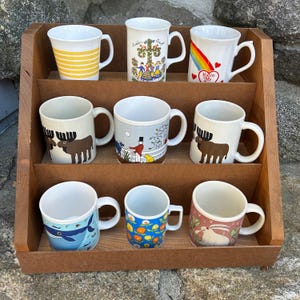 May include: A wooden display shelf holds nine ceramic mugs with various designs. The mugs feature images of a rainbow, moose, floral patterns, and other illustrations. The mugs are white with curved handles and are arranged on three shelves.