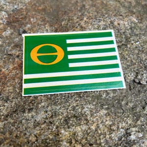 May include: A rectangular sticker with a green background and white horizontal stripes. A gold-colored symbol is on the left side. The sticker is on a rough, gray stone surface.