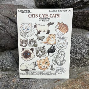 May include: A vintage cross-stitch pattern booklet titled "CATS CATS CATS!" featuring various cat designs. The cover includes the text "MINI SERIES #13 By Mary Ellen" and the alphabet. The booklet is on a stone surface.