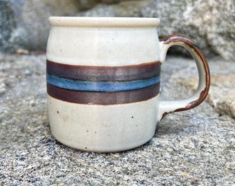 May include: A ceramic mug with a speckled off-white glaze featuring a horizontal band of dark brown and blue stripes around the center. The mug has a sturdy handle on the right side and a smooth, rounded rim, showcasing a handcrafted appearance.