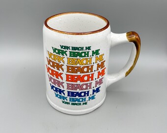 May include: White ceramic mug with a brown rim and handle. The mug features the text "YORK BEACH, ME" in a rainbow of colors. The handle has a brown gradient.
