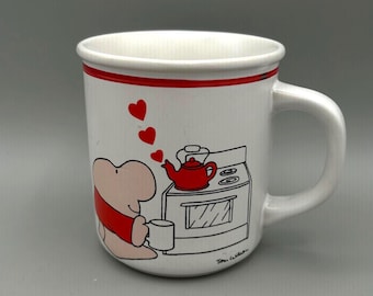 May include: White ceramic mug with a red rim and handle. The mug features a cartoon illustration of a figure in a red scarf, a red teapot, and a stove. Red hearts float above the figure. Signed "Tom Wilson".