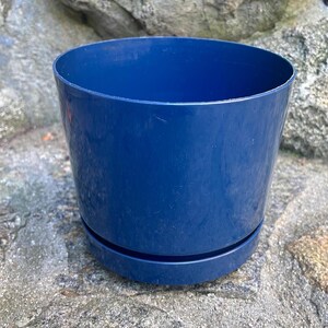May include: A solid navy blue cylindrical plant pot with a smooth, glossy finish. The container features a matching circular base tray attached at the bottom, creating a clean, uniform appearance for this functional gardening vessel.