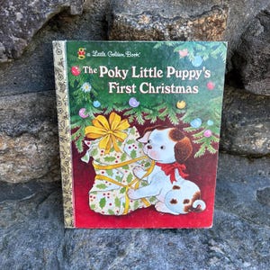 May include: A Little Golden Book titled "The Poky Little Puppy's First Christmas". The cover features a white puppy with brown spots and ears, holding a Christmas stocking with a yellow bow. The puppy is standing under a decorated Christmas tree with red, green, and gold ornaments.