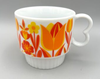 May include: A white ceramic mug featuring a vibrant floral pattern with orange, yellow, and red tulips and small flowers. The mug has a distinctively shaped heart-like handle and a ribbed base, providing a textured look to the lower section.