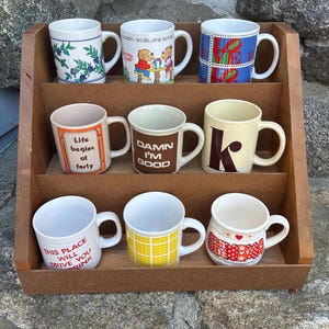 May include: A wooden display shelf holding nine ceramic mugs with various designs and text. The mugs feature floral patterns, cartoon characters, and phrases like "Life begins at forty" and "Damn I'm good."