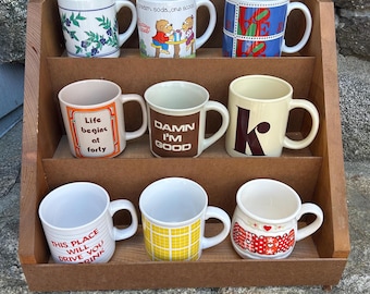 May include: A wooden display shelf holding nine ceramic mugs with various designs and text. The mugs feature floral patterns, cartoon characters, and phrases like "Life begins at forty" and "Damn I'm good."