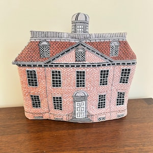 May include: A decorative brick-patterned house-shaped tea cozy with a red roof and a small cupola. The windows and door are black and white, and the cozy sits on a wooden surface.