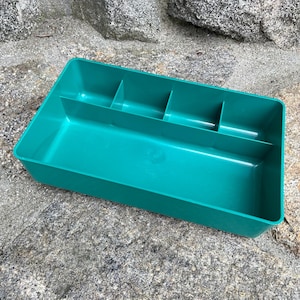 May include: A teal plastic organizer with multiple compartments. The rectangular container has a long, open section and several smaller, divided sections, ideal for storing small items. The organizer is sitting on a textured, gray stone surface.