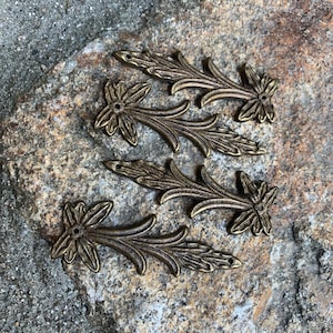 May include: Six antique gold-toned metal floral embellishments with a leaf design. The embellishments are arranged on a rough, gray stone surface.