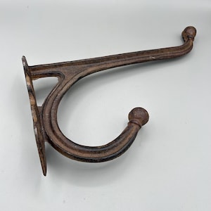 May include: A rustic, brown cast iron wall hook. The hook has a curved design with a ball at the end and a decorative support. The hook is designed to be mounted on a wall for hanging items.