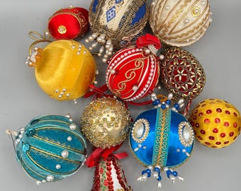 May include: A collection of twelve decorative ornaments in various shapes and colors, including spheres and a bell. The ornaments feature satin fabric, gold trim, intricate beadwork, faux pearls, and sequins in shades of blue, red, yellow, and cream.