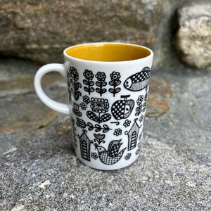 May include: A white ceramic mug with a black and white folk art design featuring houses, flowers, birds, and a fish. The mug has a yellow interior.