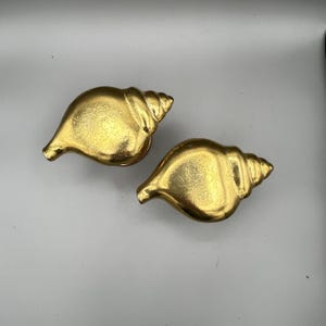 May include: Two gold-colored seashell-shaped decorative objects. The objects have a spiral design and a smooth, shiny surface. The seashells are placed on a light gray background.