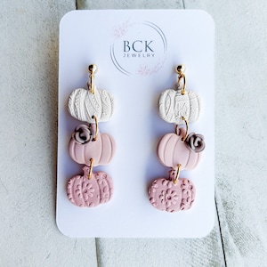 May include: A pair of dangle earrings featuring three pink and white pumpkin charms with floral details. The earrings are displayed on a white card with the text "BCK JEWELRY".