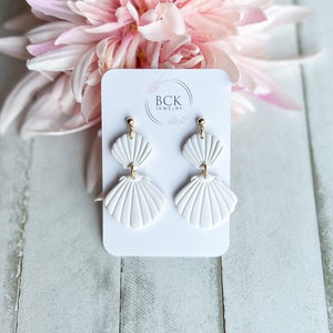 Peut inclure: Boucles d'oreilles blanches en forme de coquillage avec des détails dorés. Elles sont présentées sur une carte blanche avec le logo "BCK Jewelry". En arrière-plan, une fleur rose tendre et une surface en bois blanc.