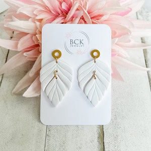 Peut inclure: Boucles d'oreilles en forme de feuille blanches avec des accents dorés. Les boucles d'oreilles sont présentées sur une carte blanche avec le texte "BCK JEWELRY".