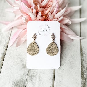 May include: A pair of beige snake skin patterned earrings with gold accents. The earrings are displayed on a white card with the text "BCK JEWELRY".