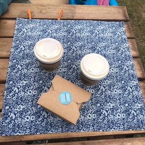 Puede incluir: Dos tazas de caf&eacute; de papel con tapas blancas y un recipiente de comida de papel marr&oacute;n sobre un mantel individual azul con estampado floral. El mantel individual tiene lazos naranjas. El recipiente de comida tiene una pegatina azul con texto blanco.