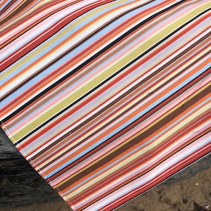 Puede incluir: Una tela a rayas multicolores con rayas diagonales rojas, naranjas, marrones, negras, blancas, azules y verdes. El borde de la tela es visible, mostrando un borde blanco con costuras rojas. Probablemente es un textil.