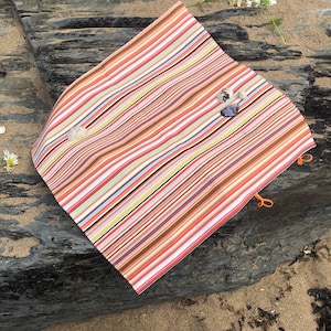 Puede incluir: Una manta de playa a rayas de colores con rayas naranjas, marrones y blancas. La manta est&aacute; doblada sobre una roca gris oscura con arena y peque&ntilde;as conchas marinas. La manta tiene lazos naranjas en las esquinas.