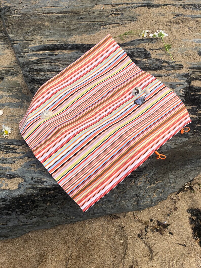 Puede incluir: Una manta de playa a rayas de colores con rayas naranjas, marrones y blancas. La manta est&aacute; doblada sobre una roca gris oscura con arena y peque&ntilde;as conchas marinas. La manta tiene lazos naranjas en las esquinas.