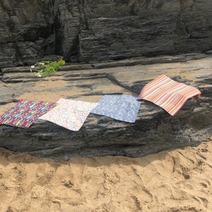 Puede incluir: Cuatro esterillas de tela coloridas se exhiben sobre una superficie rocosa cerca de la playa. Las esterillas presentan varios dise&ntilde;os, incluyendo dise&ntilde;os florales, arco&iacute;ris y a rayas. Las esterillas son rectangulares y parecen estar hechas de un material ligero.