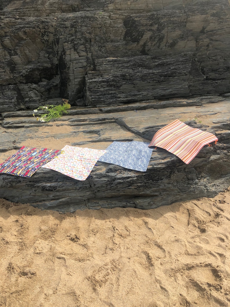 Puede incluir: Cuatro esterillas de tela coloridas se exhiben sobre una superficie rocosa cerca de la playa. Las esterillas presentan varios dise&ntilde;os, incluyendo dise&ntilde;os florales, arco&iacute;ris y a rayas. Las esterillas son rectangulares y parecen estar hechas de un material ligero.