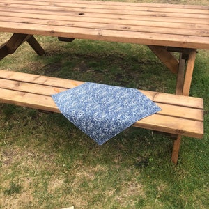 Puede incluir: Un camino de mesa con estampado floral azul y blanco se muestra sobre un banco de picnic de madera. El camino de mesa es rectangular y parece ser de tela. La mesa de picnic es de madera marr&oacute;n claro y est&aacute; al aire libre sobre c&eacute;sped.