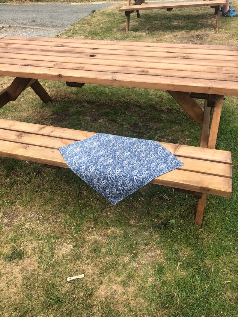 Puede incluir: Un camino de mesa con estampado floral azul y blanco se muestra sobre un banco de picnic de madera. El camino de mesa es rectangular y parece ser de tela. La mesa de picnic es de madera marr&oacute;n claro y est&aacute; al aire libre sobre c&eacute;sped.