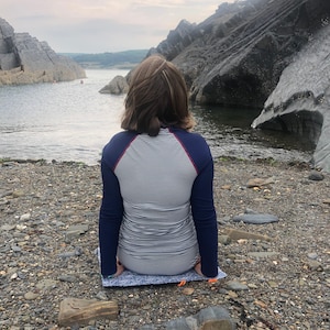 Puede incluir: Una persona con una camisa de manga larga a rayas azul marino y blanco est&aacute; sentada en una esterilla estampada en una playa rocosa. La persona mira hacia el agua, con grandes formaciones rocosas a ambos lados.