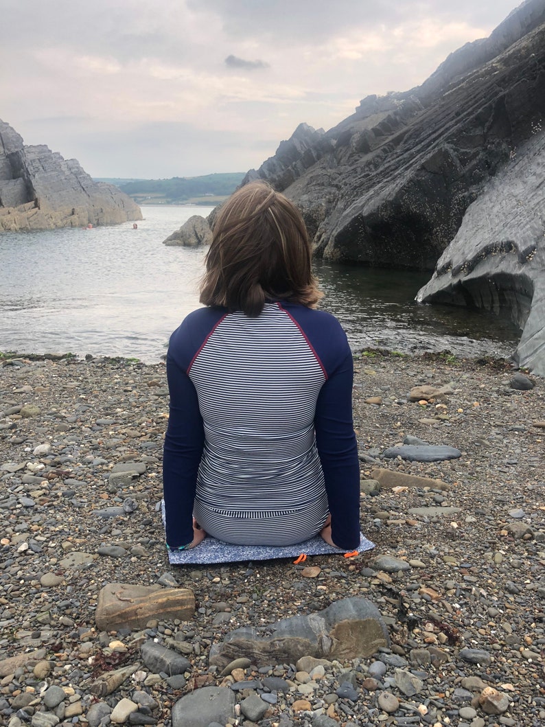 Puede incluir: Una persona con una camisa de manga larga a rayas azul marino y blanco est&aacute; sentada en una esterilla estampada en una playa rocosa. La persona mira hacia el agua, con grandes formaciones rocosas a ambos lados.