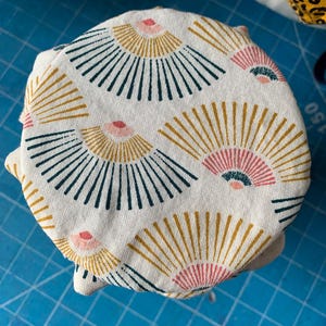 Puede incluir: Una pila de objetos cubiertos de tela, posiblemente recipientes o tapas, con un patrón repetitivo en forma de abanico. El diseño incluye elementos azul marino, dorados y rosas sobre un fondo crema. Los objetos están apilados sobre una superficie azul.