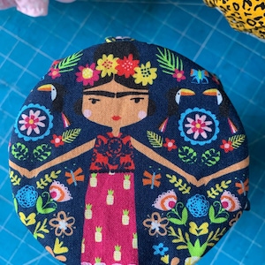 Puede incluir: Un objeto redondo de tela azul marino con una ilustración estilizada de una mujer adornada con una corona floral. El diseño incluye tucanes, flores y un vestido rosa con motivos de piña. El objeto está rodeado de coloridos patrones florales y de hojas.