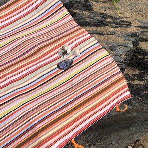 Puede incluir: Una manta de playa a rayas de colores con rayas naranjas, marrones, rojas y azules. La manta tiene lazos naranjas en las esquinas. Conchas marinas y una flor est&aacute;n en la arena junto a la manta.