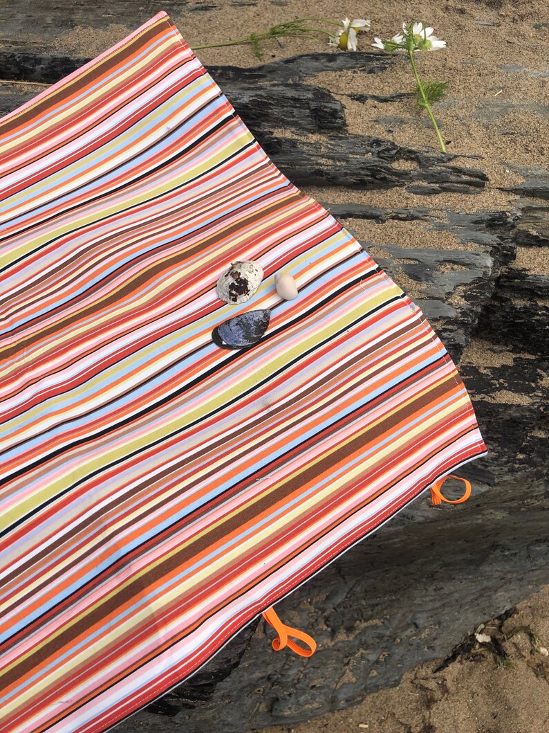 Puede incluir: Una manta de playa a rayas de colores con rayas naranjas, marrones, rojas y azules. La manta tiene lazos naranjas en las esquinas. Conchas marinas y una flor est&aacute;n en la arena junto a la manta.