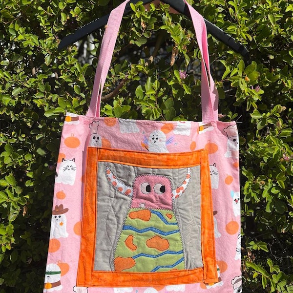 Puede incluir: Bolso tote rosa con un estampado de Halloween de fantasmas, calabazas y gatos. El bolso presenta un parche central con un diseño de monstruo en rosa, gris, naranja y verde. El bolso tiene asas rosas.