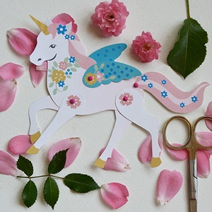 May include: A white paper unicorn with pink, blue, and yellow floral details. The unicorn has a gold horn, blue wings, and a pink tail. The unicorn is surrounded by pink rose petals and green leaves.