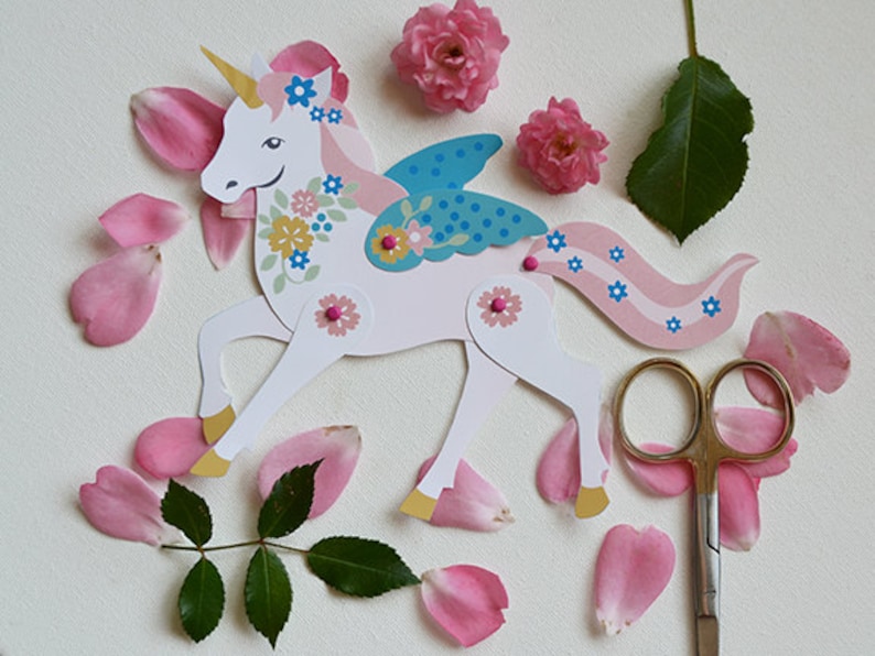 May include: A white paper unicorn with pink, blue, and yellow floral details. The unicorn has a gold horn, blue wings, and a pink tail. The unicorn is surrounded by pink rose petals and green leaves.