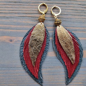 Puede incluir: Un par de pendientes colgantes con plumas de cuero doradas, rojas y azules. Los pendientes tienen aros dorados y cuentas doradas.