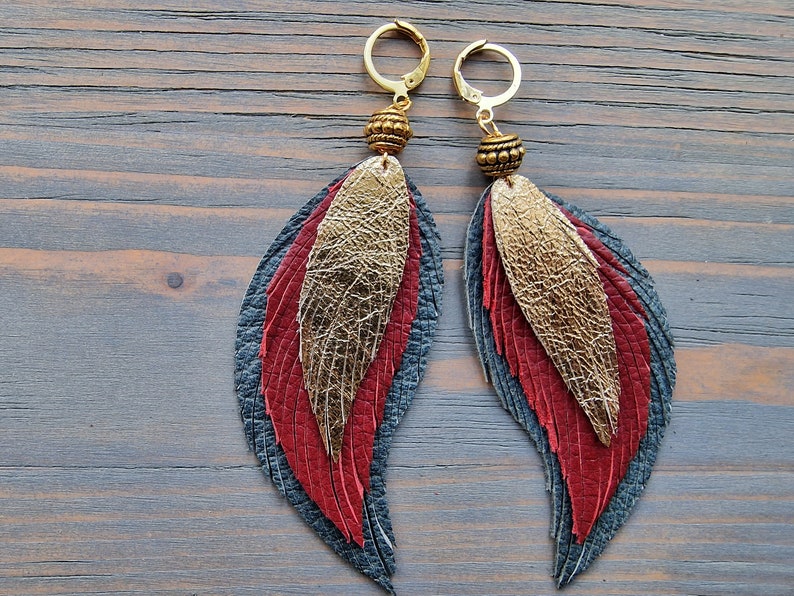 Puede incluir: Un par de pendientes colgantes con plumas de cuero doradas, rojas y azules. Los pendientes tienen aros dorados y cuentas doradas.