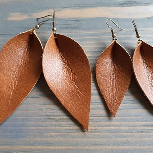 May include: Four brown leather leaf earrings with bronze-colored ear wires are displayed against a gray-blue wooden background. The earrings have a textured surface and a natural, organic shape, suggesting a nature-inspired theme.