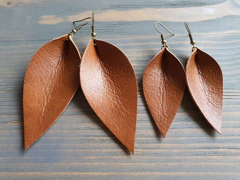 May include: Four brown leather leaf earrings with bronze-colored ear wires are displayed against a gray-blue wooden background. The earrings have a textured surface and a natural, organic shape, suggesting a nature-inspired theme.
