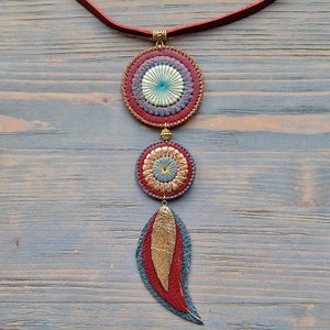 Puede incluir: Un collar de cuero rojo con tres colgantes circulares. Los colgantes est&aacute;n decorados con c&iacute;rculos conc&eacute;ntricos de cuero rojo, dorado y azul. El collar termina con un dije de pluma de cuero rojo.