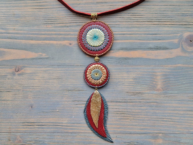 Puede incluir: Un collar de cuero rojo con tres colgantes circulares. Los colgantes est&aacute;n decorados con c&iacute;rculos conc&eacute;ntricos de cuero rojo, dorado y azul. El collar termina con un dije de pluma de cuero rojo.