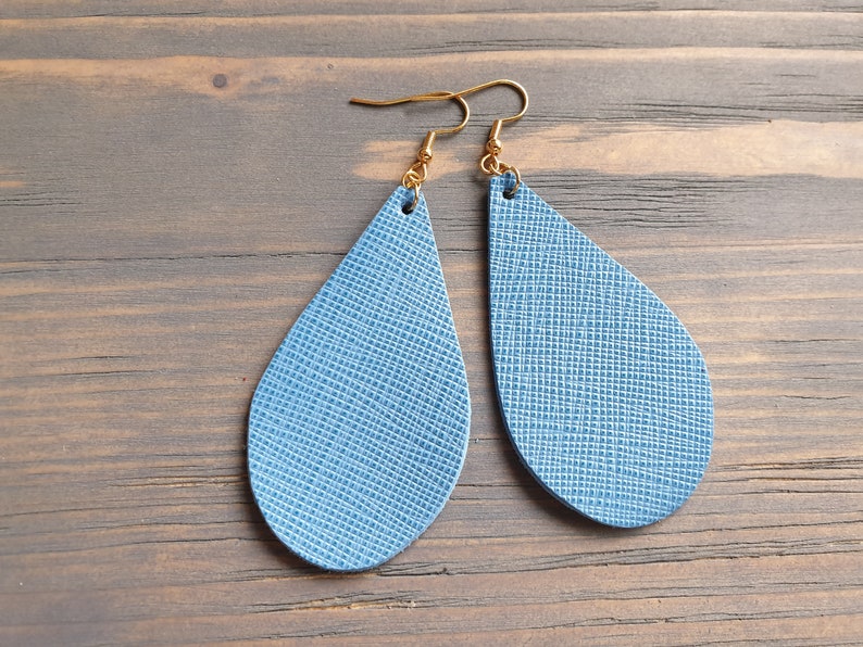 May include: A pair of teardrop-shaped earrings made of blue leather with a textured surface. The earrings have gold hooks.