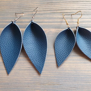 May include: Two pairs of navy blue leather leaf earrings. One pair has silver-colored ear wires, and the other has gold-colored ear wires. The earrings have a textured surface and a teardrop shape, resembling leaves.