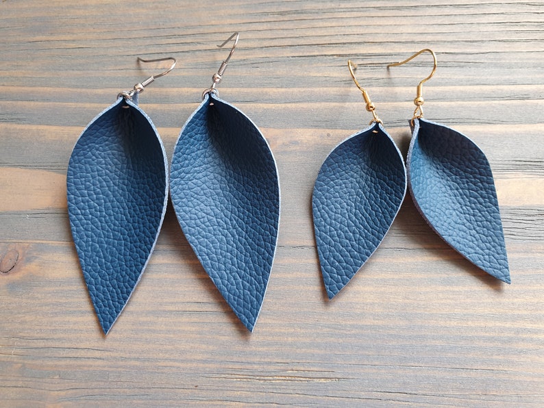 May include: Two pairs of navy blue leather leaf earrings. One pair has silver-colored ear wires, and the other has gold-colored ear wires. The earrings have a textured surface and a teardrop shape, resembling leaves.