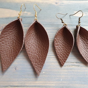May include: Four brown leather leaf earrings with gold and bronze-colored hooks. The earrings have a textured surface and a teardrop shape. Two pairs of earrings are displayed on a blue and gray wooden surface.
