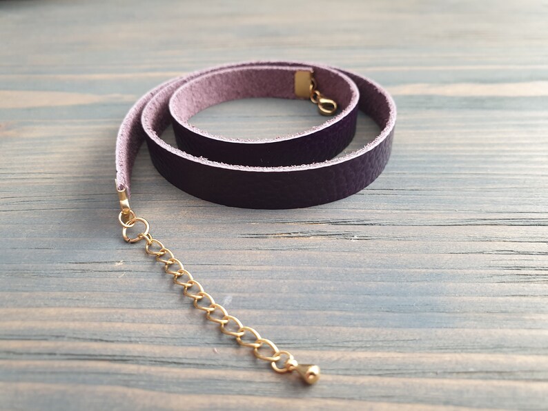 May include: A purple leather choker necklace with a gold chain extender. The choker is rolled and has a smooth, textured surface.